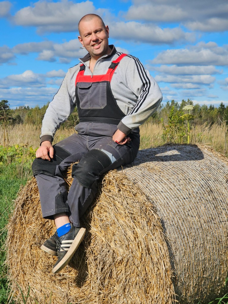 Андрей Пальченко в рабочем комбинезоне на тюке сена, поле